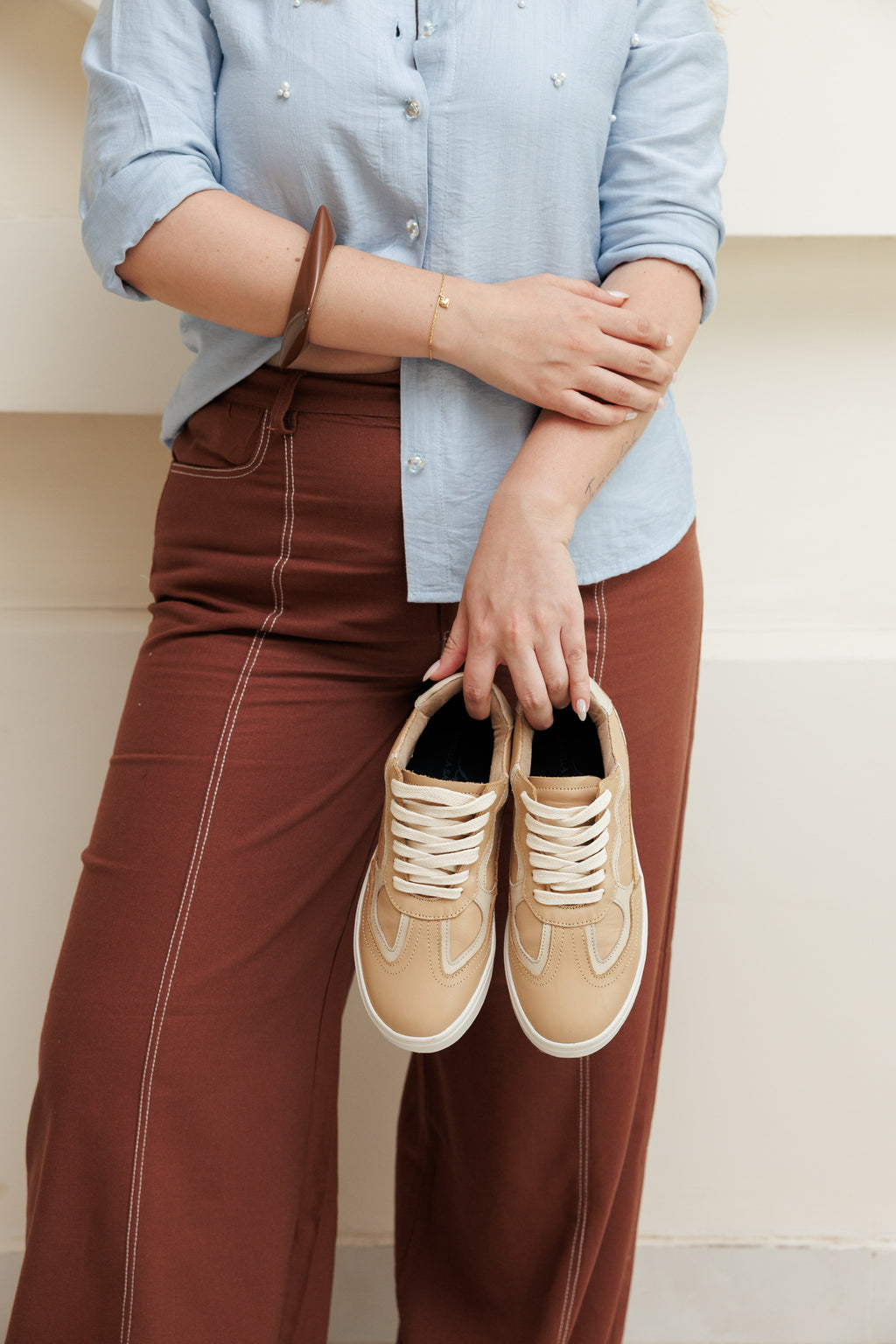 Tenis Athenas para mujer en cuero color beige, diseño minimalista con suela blanca spanson y cordones a tono, sostenidos por una modelo.